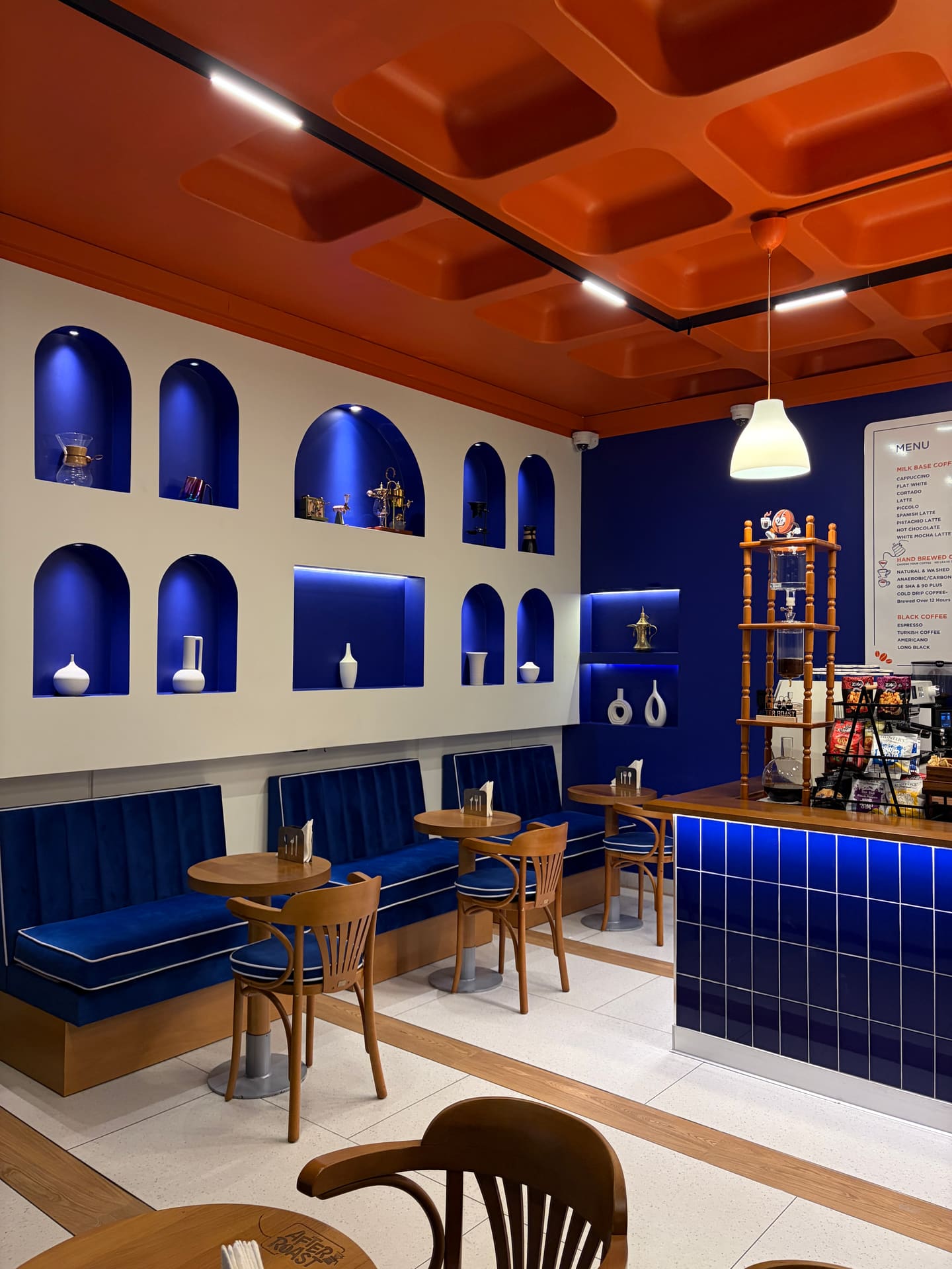 Blue velvet banquette seating and round wooden tables beside a wall of lit arched niches holding ceramics and brewing gear.