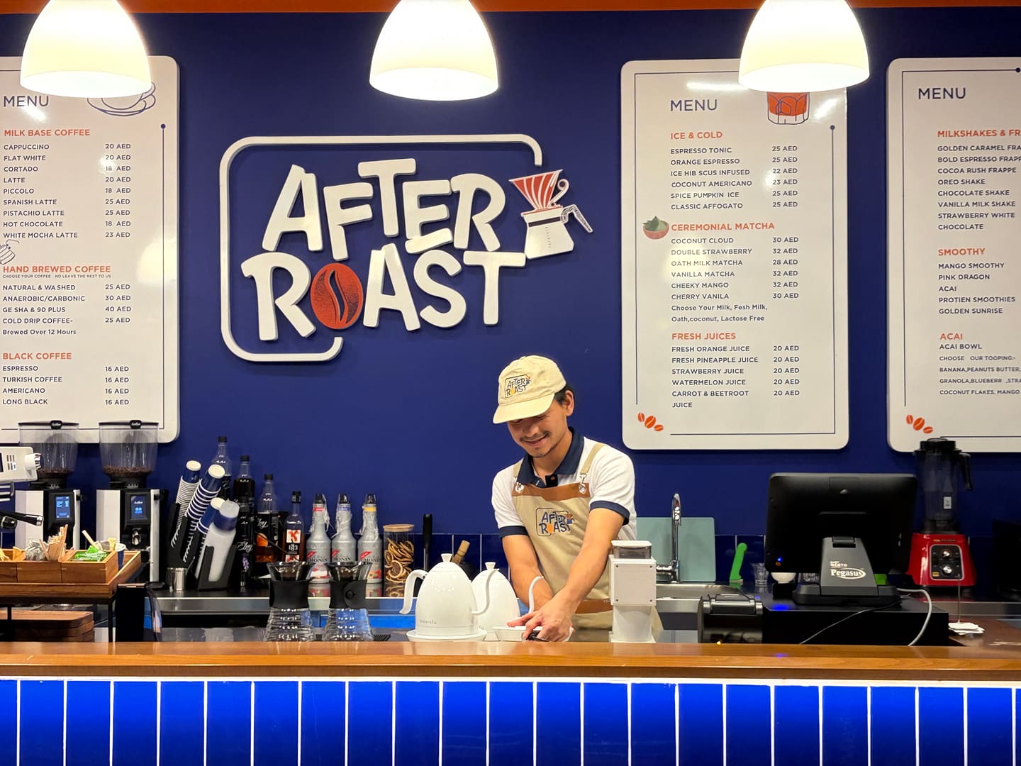A barista in an After Roast cap and apron setting down a white gooseneck kettle at the counter, the coffee menu boards on the cobalt wall behind him.