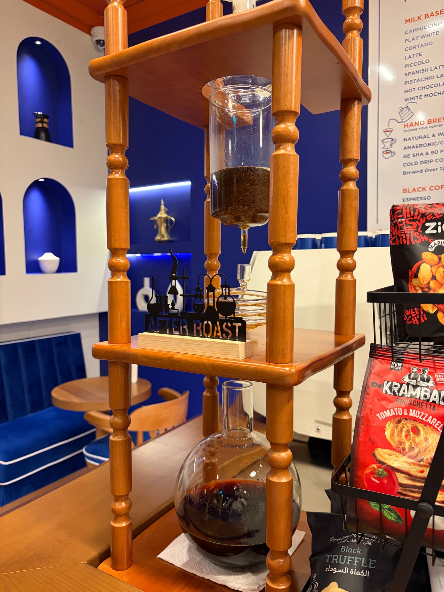 A glass cold-drip tower on a turned wooden stand, dark coffee collecting in the round carafe below.