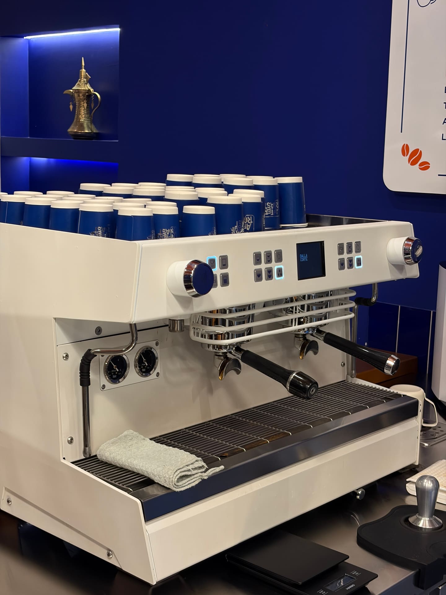 A white Dalla Corte espresso machine on the bar, stacked blue After Roast takeaway cups warming on top.