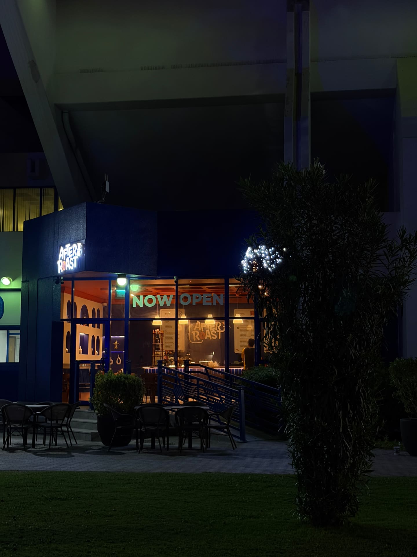 The After Roast corner unit seen at night from across the lawn, outdoor tables and chairs set out under the illuminated sign.